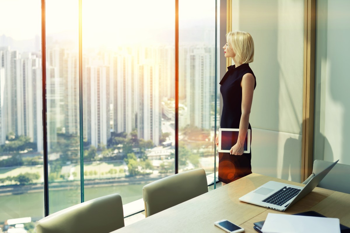 Woman in black dress standing in business office with iPad in her hand watching out of the window over downtown Hong Kong